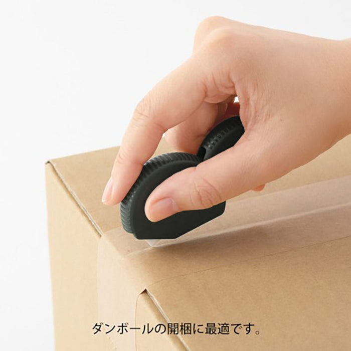 Hand using a black box-cutting carton opener on a cardboard box with Japanese text at the bottom.
