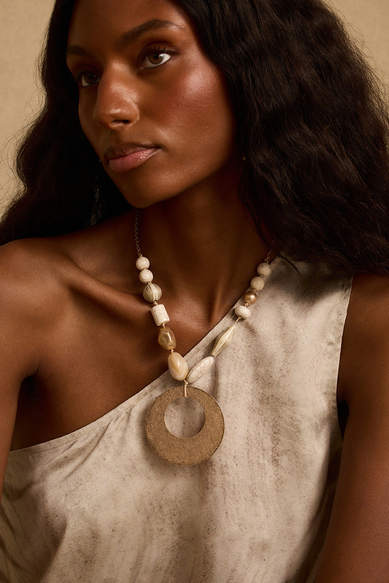 Beaded statement necklace with gold ring pendant worn on-model over a one-shoulder top.