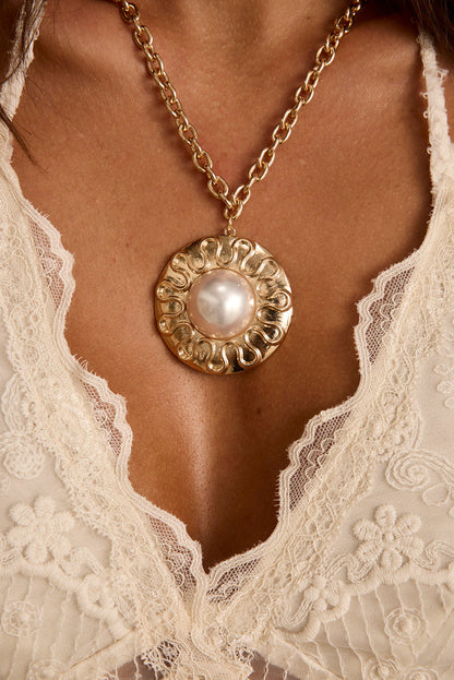 Gold pendant necklace with a central pearl, worn over a white lace top.