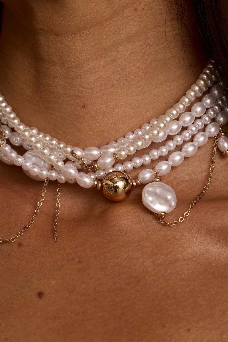 Layered pearl choker with gold ball pendant and baroque pearl drop, worn on neck.