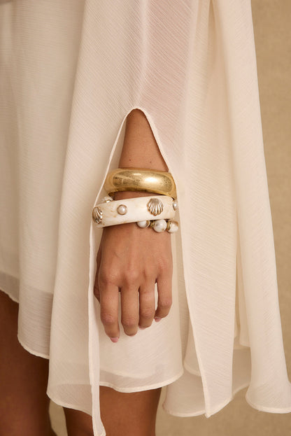 Ivory chiffon cape dress sleeve draping over a wrist stacked with gold shell bangles.