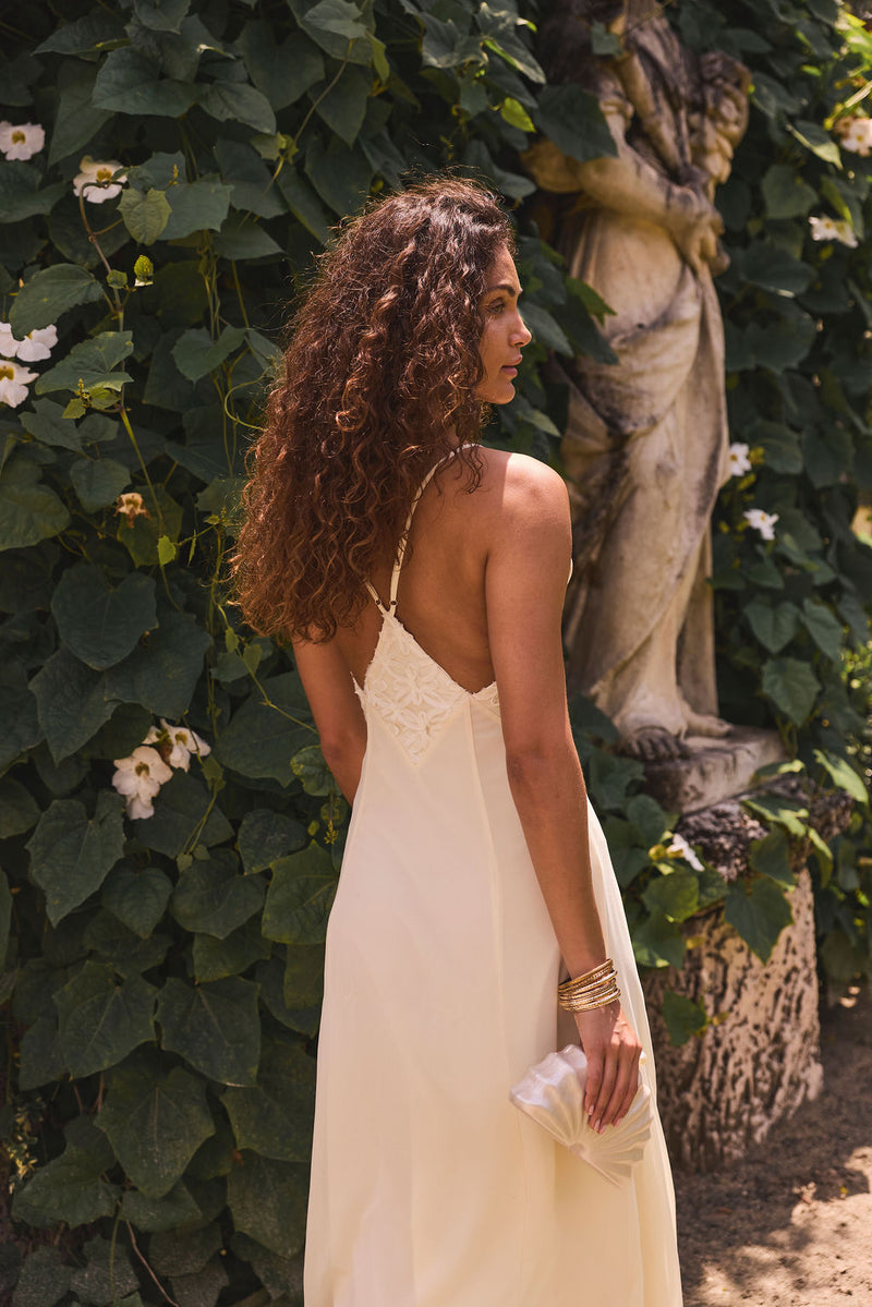 Back view of cream chiffon maxi dress with embroidered waist and crossed straps in a garden.