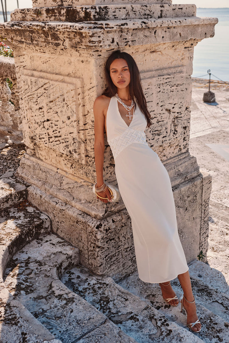 Model wearing the 12th Tribe Penelope White Halter Midi Dress, an elegant white midi dress featuring a halter top, beautiful floral embroidered waist design, and an adjustable halter tie back closure. The perfect add to your white maxi dresses line-up for beach vacation vibes, resort wear, and summer nights out.