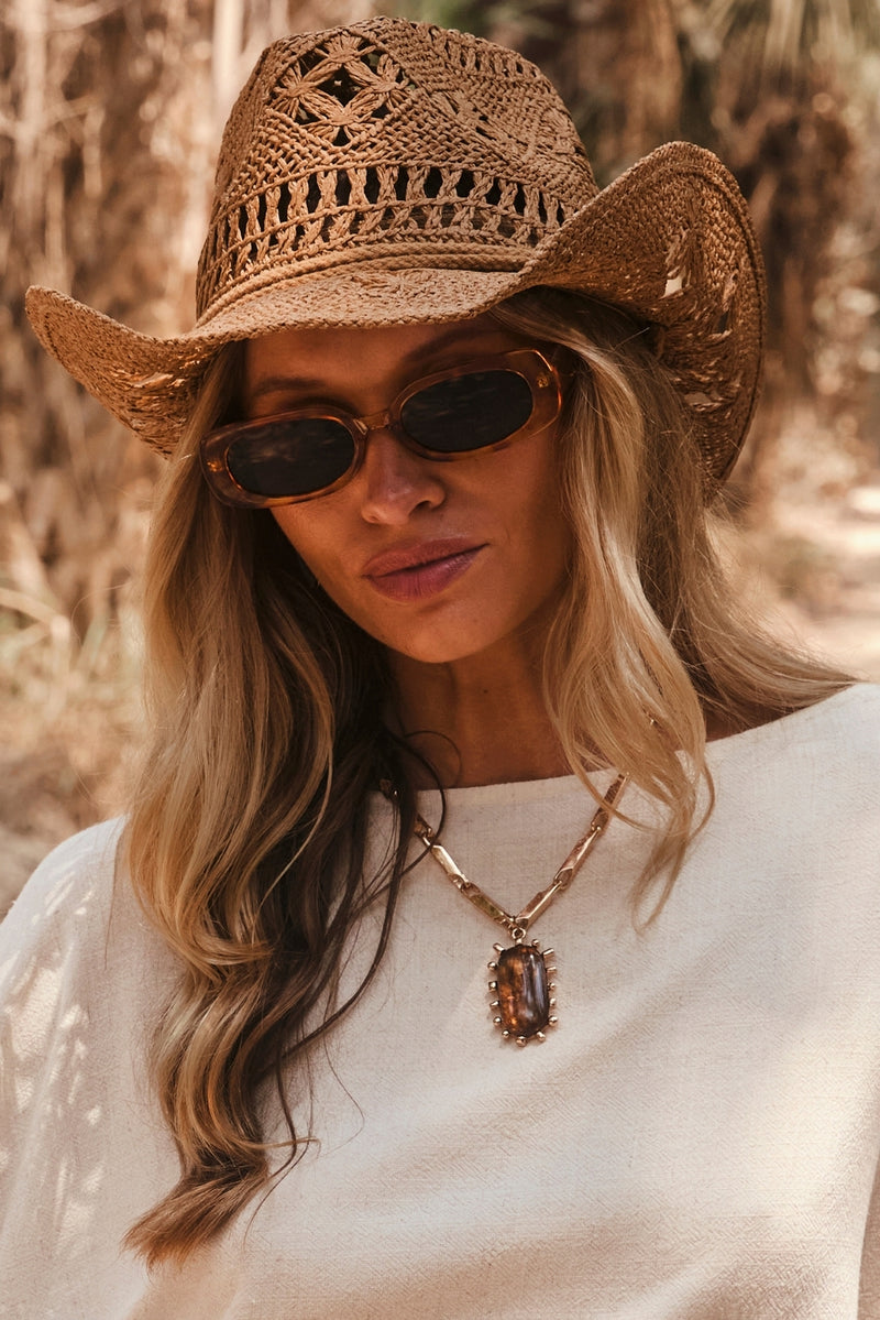 Gold statement necklace with amber stone pendant styled with straw cowboy hat and white top.