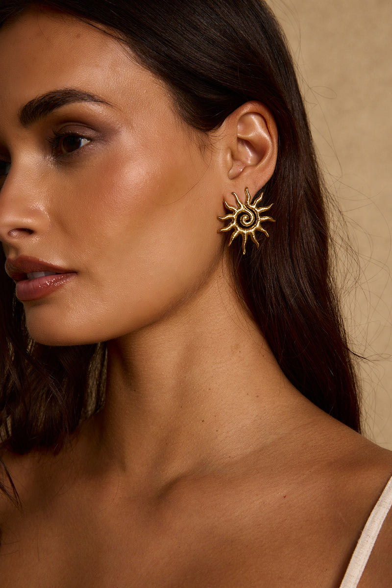 Textured gold sunburst stud earring with spiral center, worn on-model against a tan backdrop.