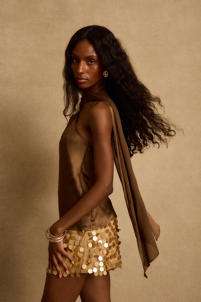 Side profile of olive satin scarf top with flowing tie-neck and gold paillette mini skirt.